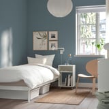 Minimalist bedroom with white VEVELSTAD bed, blue walls, natural rug, wooden chair, side table with books, lamp, and plants near window.