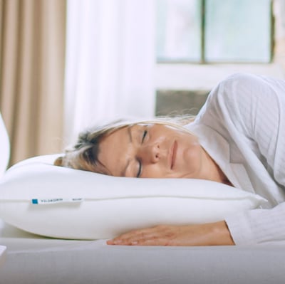 A woman sleeps peacefully on a white VILDKORN pillow designed for stomach sleepers, made from recycled polyester and cotton.
