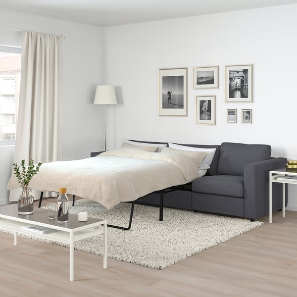 Modern grey sofa-bed in minimalist bedroom with white decor, rug, coffee table, photos, lamp, and curtains.