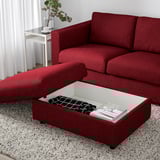 Red sofa with ottoman, lift-up seat revealing storage. On rug, near side table and plant.