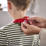 Child wearing green and white striped apron, adult fastening red neck strap, likely preparing for cooking or baking activity.