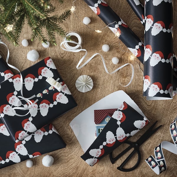 Gift wrapping scene: black wrapping paper with santa faces, scissors, ribbon, and decorations.