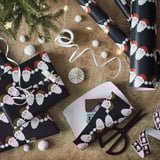 Gift wrapping scene: black wrapping paper with santa faces, scissors, ribbon, and decorations.