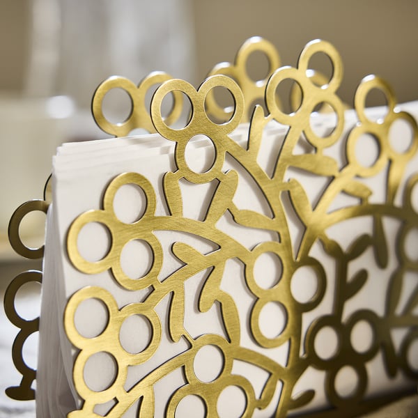 Golden steel napkin holder, intricate circles and lines design, holding white napkins, reflecting light.