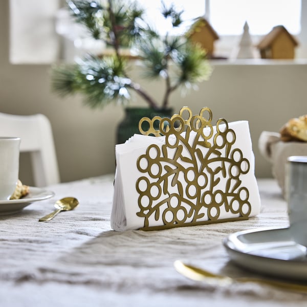 Gold VINTERFINT napkin holder with circular cutouts stands near white plates and cups on a table, smaller than the plates.