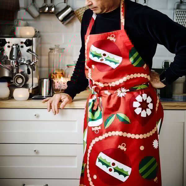 Person in festive red apron stands in kitchen. Apron has green and white ornaments, larger than standard kitchen items.