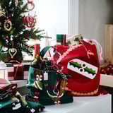 Christmas-themed VINTERFINT fabric bag, red with green trim, holding wine bottles next to decorated tree. Larger than nearby gifts.