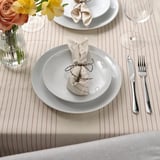Table set with white plates, folded napkins, silverware, wine glass, and striped tablecloth; flowers in vase.