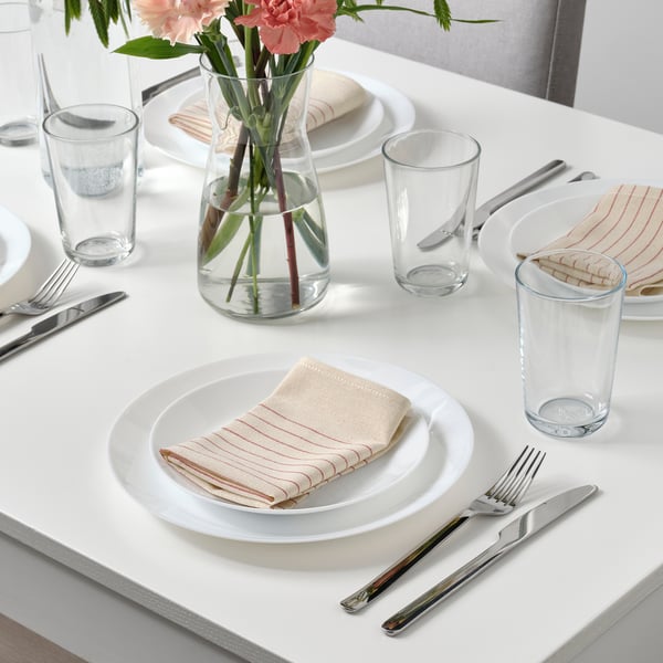 Table setting with white plates, folded striped napkins, glasses, cutlery, and a vase with flowers, arranged neatly on a white table.