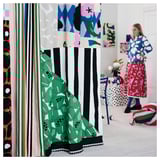 Vibrant room with colorful abstract curtains & patterns. Woman in blue & white polka dot outfit stands near.