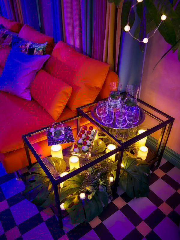 Bright, colorful living room with a red couch, pillows, and checkered floor. Glass table holds candles, cups, and a gift box. Modern, vibrant decor.