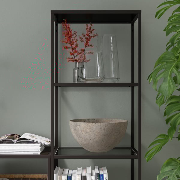 Modern bookcase with glass shelves holding books, a stone bowl, and vases with red flowers. A plant is partially visible.
