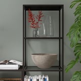 Modern bookcase with glass shelves holding books, a stone bowl, and vases with red flowers. A plant is partially visible.