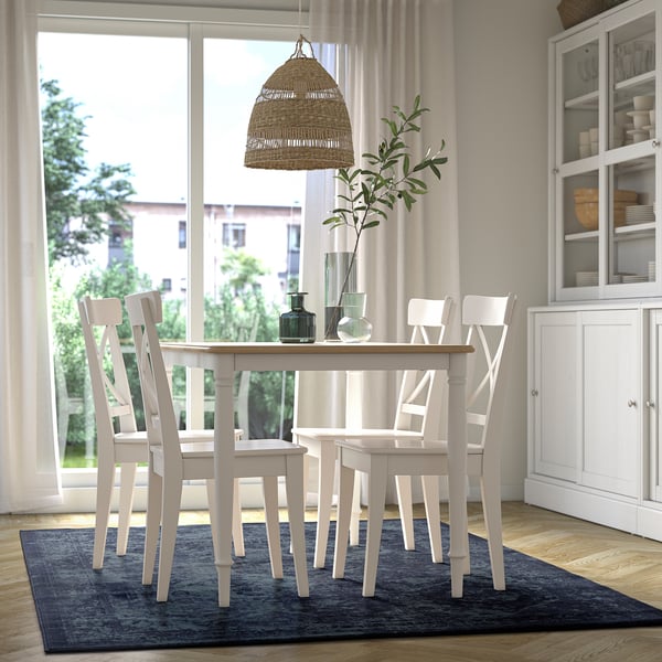 Bright dining area with white VONSBÄK chairs, a small table, dark blue rug, hanging lamp, and a glass cabinet. Large window shows buildings and trees.
