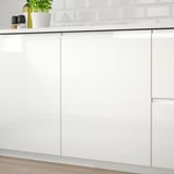 Modern kitchen with white high-gloss VOXTORP cabinets, featuring integrated handles. A potted plant and a bottle rest on the countertop.