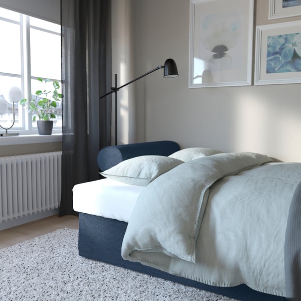 Bed by window with plant, lamp, and art. Soft, light blue bedding on comfortable sofa-bed with storage.