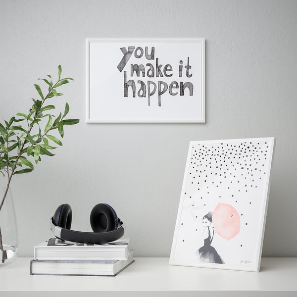 Minimal desk setup with art print you make it happen, stacked books, headphones, plant, and leaning art frame.