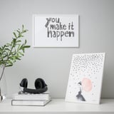 Minimal desk setup with art print you make it happen, stacked books, headphones, plant, and leaning art frame.