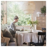 Frau liest am Tisch mit ADLAD Potpourri, Kerze, Vase. Natürliches Licht füllt den Raum mit Holzdekor und Pflanzen.