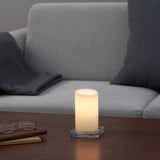 Zylindrische LED-Kerze auf Glassockel auf Holztisch, beleuchtetes warmweißes Licht, hinter grauer gepolsterter Couch mit Decke.