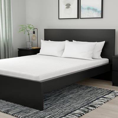 Modernes Schlafzimmer mit schwarzer Queensize-Matratze, weißen Kissen, Nachttisch, Lampe, Pflanze, Kunst und gestreiftem Teppich.