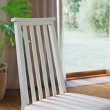 Gemütliche Indoor-Szenerie: weißer Lattenstuhl am Fenster, üppige Pflanze, brauner Vorhang, heller Holzboden.