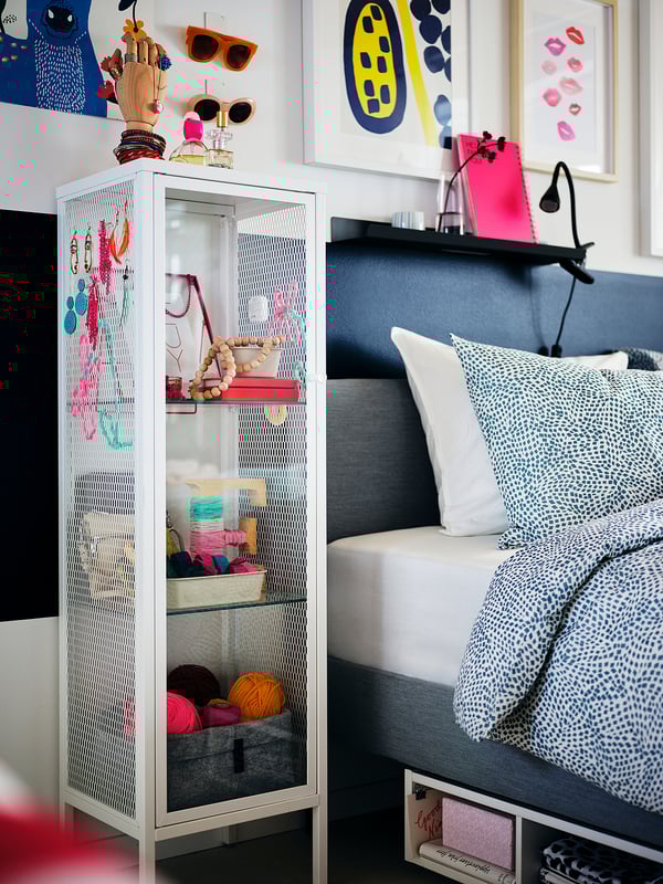 Moderne Schlafzimmerecke mit weißem BAGGEBO-Aufbewahrungsschrank mit bunten Accessoires. Graues Bett mit großen Kissen, lebendige Kunstrahmen an der Wand.