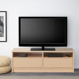 Ein hölzerner TV-Ständer mit einem Flachbildfernseher darauf, einem Stapel Bücher, einer Blackbox und einer Fernbedienung. Der Ständer verfügt über Regale und Schubladen.