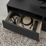 Offener schwarzer TV-Ständer, in dem Kopfhörer, Kabel, Fernbedienungen und ein Gerät zu sehen sind. Organisiert Elektronik und Zubehör.