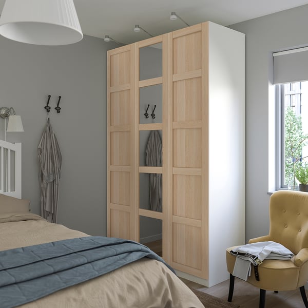 Schlafzimmer mit BERGSBO-Kleiderschrank aus hellem Holz, großem Spiegel, hängenden Kleidungsstücken, Bett mit grauer Bettwäsche, Wandlampe, gelbem Stuhl und Fenster.