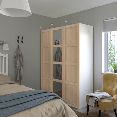 Modernes Schlafzimmer mit großem BERGSBO-Holzschrank und Spiegeltür, positioniert zwischen Bett und Fenster. Graue Wände, gelber Stuhl und hellbraune Bettwäsche.