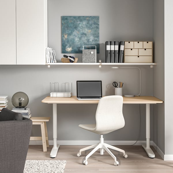 Modernes Homeoffice mit hellem Holzschreibtisch und weißem Stuhl, Laptop, Büchern und BERGSHULT Wandregal darüber. Graue Wände.