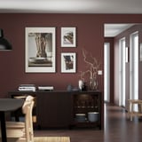 BESTÅ Sideboard aus dunklem Holz an burgunderroter Wand mit Kunstwerk. Es ist breit, fast vom Boden bis zur Decke, mit stapelbarem Geschirr und dekorativen Vasen.