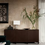 Modernes Wohnzimmer mit braunem BESTÅ-Stauraum, weißer Lampe, hoher Pflanze in klarer Vase, grünem Stuhl, abstrakter Wandkunst.