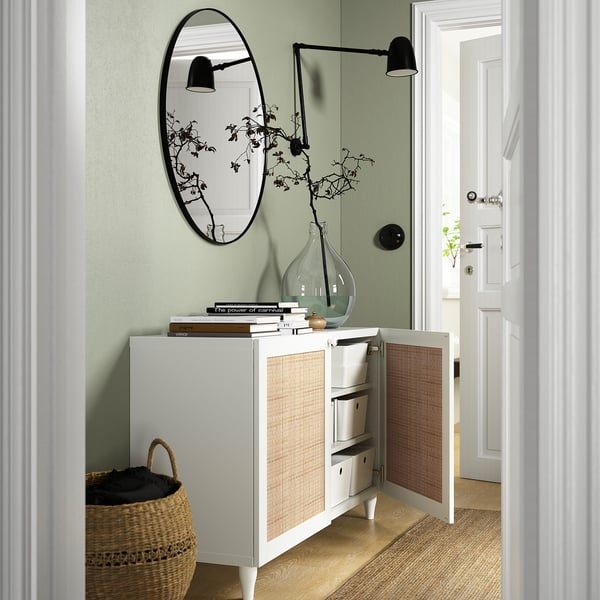 Weißes BESTÅ Sideboard mit offenen Türen, in dem Aufbewahrungsboxen, Dekorationsartikel und Bücher ausgestellt sind. Runder Spiegel oben, flankiert von modernen Lampen.