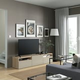Modernes Wohnzimmer mit BESTÅ-TV-Bank, TV, Kunstdrucken, Stehlampe, Couchtisch aus Glas, grauen Wänden und großem Fenster.
