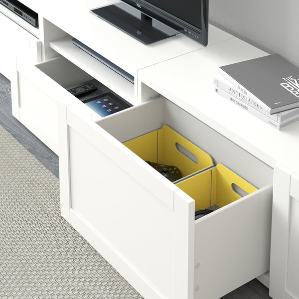 Weißer BESTÅ TV-Schrank mit verstellbaren Regalen, integriertem Kabelmanagement und gelber Schublade mit Gamecontrollern.