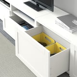 Weißer BESTÅ TV-Schrank mit verstellbaren Regalen, integriertem Kabelmanagement und gelber Schublade mit Gamecontrollern.