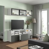 Modernes Wohnzimmer mit weißer und grauer BESTÅ-TV-Bank, Flachbildfernseher, Glasregalen und Vorhängen.