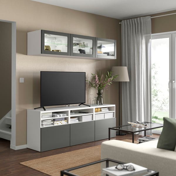 Modernes Wohnzimmer mit weiß-grauem BESTÅ-TV-Ständer, Flachbildfernseher, Wandschränken, Glastürdisplay, grauen Vorhängen, Holzboden und beigem Sofa.
