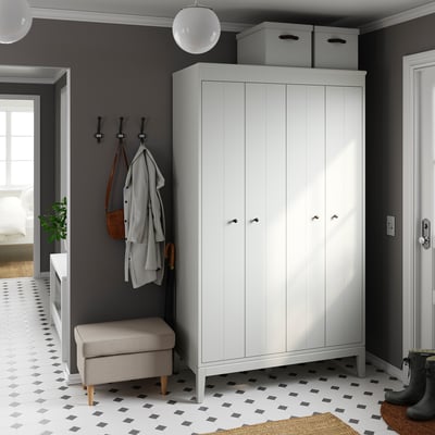 Kleines Zimmer mit grauen Wänden, weißem Kleiderschrank, schwarz-weißem Fliesenboden. Graue Bank, Kleiderhaken und hängende Mäntel, Taschen.