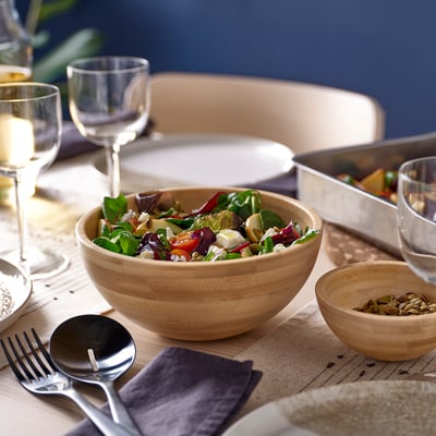 Große Holzschüssel mit Salat auf dem Tisch. In der Nähe von Weingläsern, Tellern und einer kleineren Schüssel mit Samen. Begleitet von Servierbesteck und Servietten.