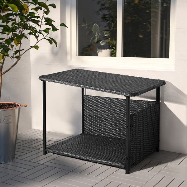 Outdoor-Tisch aus Korbgeflecht, dunkelgrau, rechteckig mit Metallbeinen, in der Nähe von Fenster und Mülleimer.