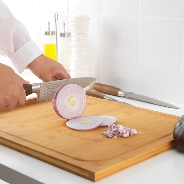 Eine Person schneidet mit einem großen, scharfen Messer eine Zwiebel auf einem hölzernen Schneidebrett in Scheiben. Fein gehackte Zwiebelstücke liegen bereits auf dem Brett.