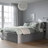 Graues BRIMNES Bett mit Schubladen und Kopfteil, hellblauen Wänden, großem Fenster und Hängelampe. Ideal für kleine Räume.