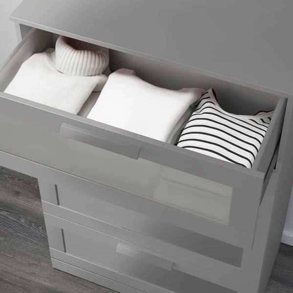Graue Kommode mit oberer Schublade aus Milchglas, teilweise geöffnet, die den Blick auf ordentlich gefaltete Kleidung freigibt.