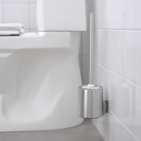 Bürstenhalter aus Metall, der an einer gefliesten Wand befestigt ist und eine weiße Bürste hält, neben einem weißen Waschbecken. Teil der BROGRUND-Serie für aufgeräumtes Bad.