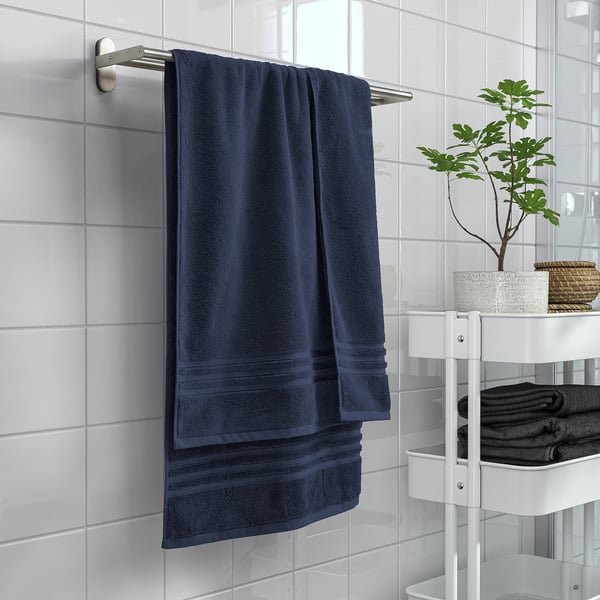 Blaues BROKGLIM Handtuch auf rostfreiem Stab, neben weißem Regal mit gefalteten Handtüchern und Pflanze.