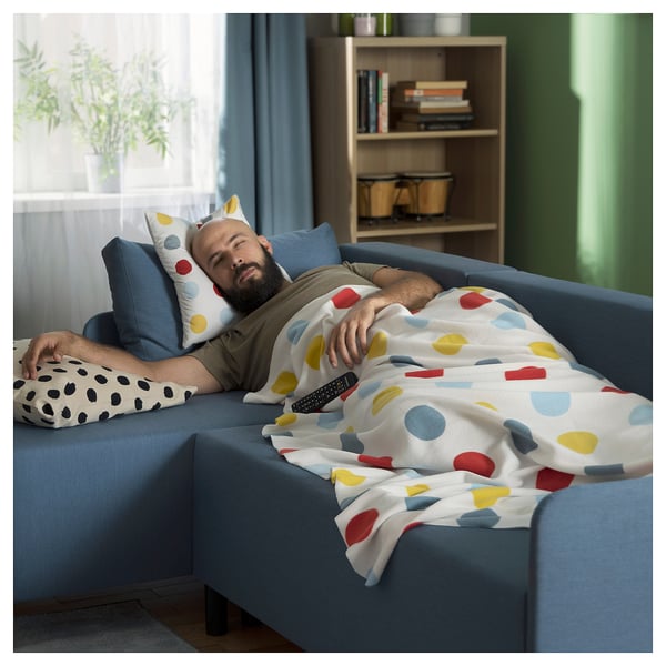 Ein Mann schläft auf einem blauen BRUKSVARA-Sofa, das mit einer weißen Decke mit bunten Flecken bedeckt ist. Der Raum verfügt über Holzregale mit Büchern und Pflanzen.
