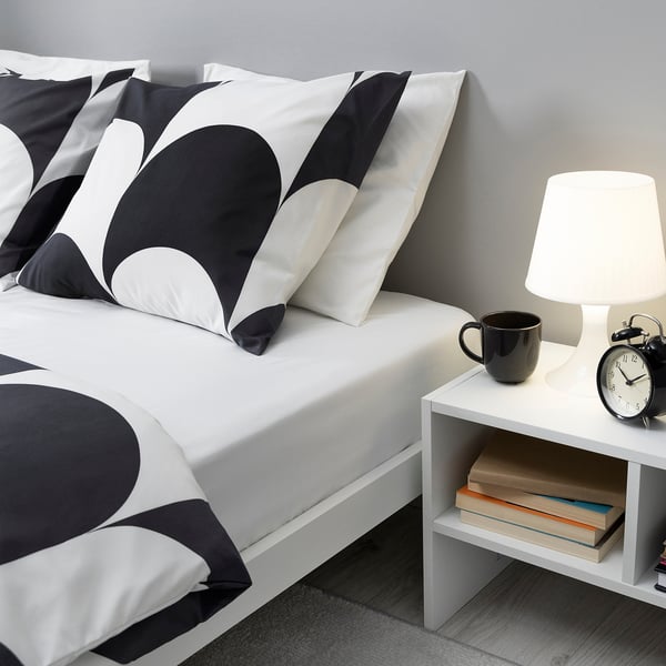 Bett mit BRUKSVARA schwarz/weißen Bogenkissen, weißer Lampe, Becher, Uhr und Büchern auf dem Nachttisch.